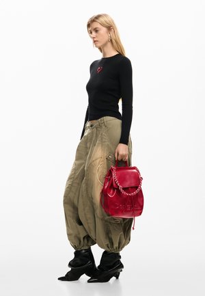 Plavuša u crnoj majici s dugim rukavima s crvenim srcem, širokim maslinastim hlačama, crnim čizmama, drži crvenu kožnu torbu na bijeloj pozadini.