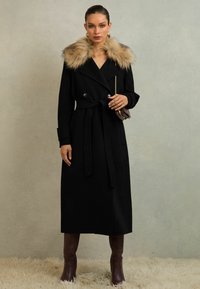 Melnā divrindu mētelis ar bežīgu mākslīgās kažokādas apkakli, jostu viduklī un sānu kabatām, savienots ar ceļa augstuma brūnajām zābakiem.