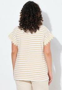 Bílý pruhovaný top se žlutými horizontálními pruhy a krajkovými detaily na rukávech. Volný střih se rovnými linemi a jemnou texturou tkaniny.
