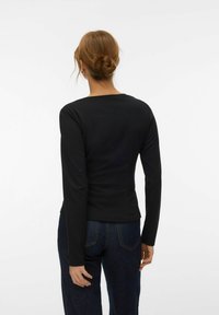 Femme aux cheveux roux attachés en chignon, portant un haut à manches longues noir et un jean bleu foncé, vue de dos sur un fond blanc.