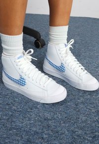 Visoke bele superge iz gladkega usnja z modrimi karirastimi poudarki na swoosh-u in režah za vezalke, v combinatie z belimi nogavicami do gležnja.