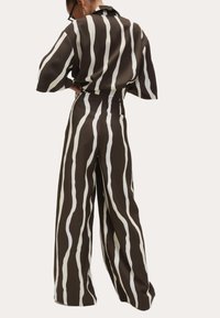 Femme portant un pantalon large à rayures verticales noir et blanc et une chemise oversized à manches courtes assortie, se tenant avec les mains sur les hanches, vue de dos.