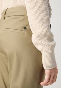 Gros plan d'une personne portant un pantalon beige avec un bouton sur la poche arrière et une manche de pull en tricot crème visible.