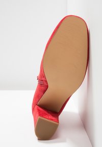 Chaussure à talon haut en daim rouge avec un bout arrondi et une semelle texturée. Le talon est épais et la tige présente un design sans couture.