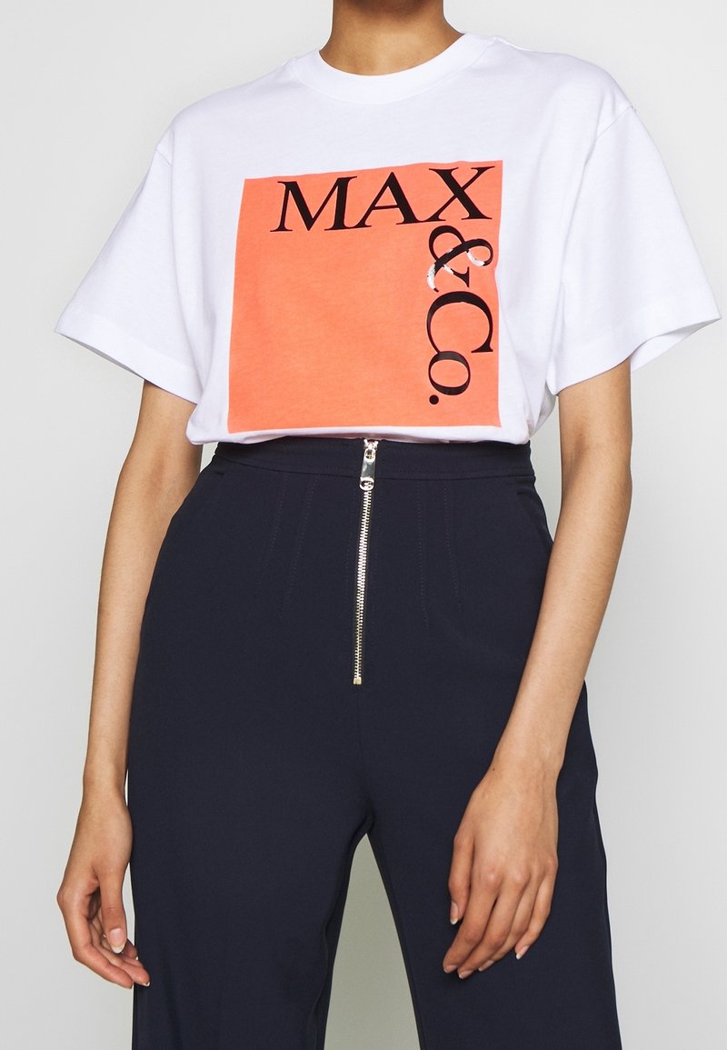Personne portant un T-shirt blanc avec un grand carré orange et un texte noir "MAX & Co.", associé à un pantalon taille haute bleu marine avec une fermeture éclair à l’avant.