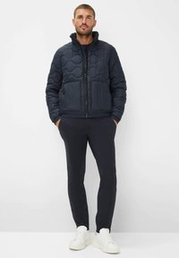 Giacca imbottita blu navy con colletto alto, due tasche frontali e polsini elasticizzati, abbinata a pantaloni scuri e sneakers bianche. Texture del tessuto liscia.