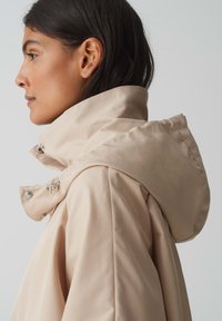 Giacca impermeabile beige chiaro con colletto alto e cappuccio staccabile, dotata di dettagli in metallo argentato e una superficie liscia e opaca.