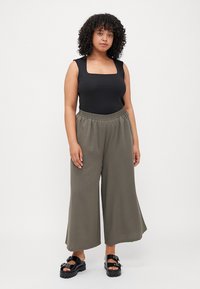 Μαύρο αμάνικο bodysuit συνδυασμένο με χαλαρές, ελαιόχρωμες παντελόνες σε άνοιγμα. Μοντέλο που φοράει χοντρές μαύρες σαγιονάρες με διακοσμητικά στοιχεία.