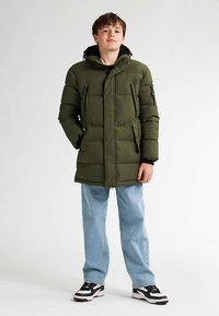 Zelená prošívaná bunda s kapucí, předním zipem, dvěma bočními kapsami a žebrovanými manžetami, nošená přes černé tričko a světle modré džíny.