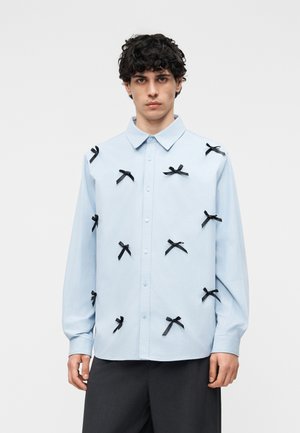 Fiatal férfi világoskék, több kis fekete masnival díszített gombos ingben, sötét nadrággal párosítva.