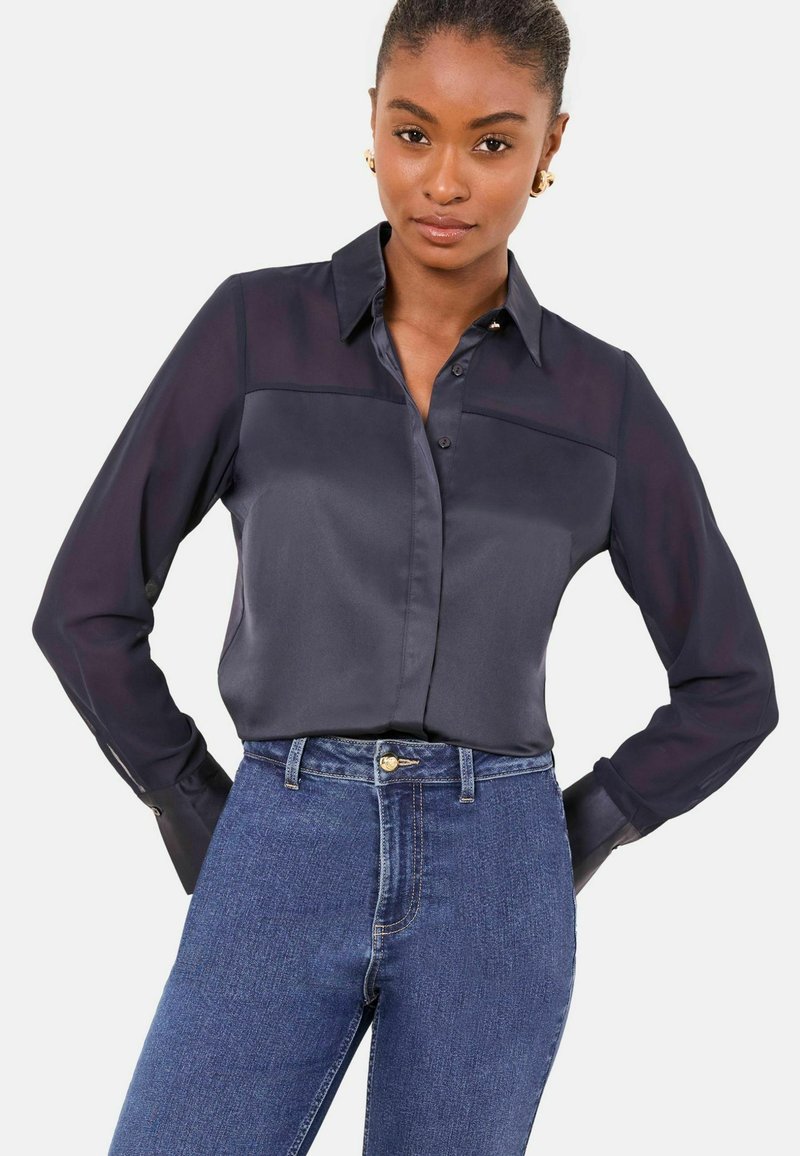 Cămașă bleumarin cu nasturi, cu finisaj satinat, mâneci lungi translucide contrastante și guler ascuțit, combinată cu blugi albastru clasic.