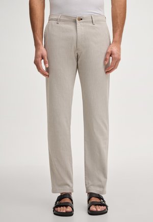 Mężczyzna w beżowych prostych spodniach i czarnych sandałach z klamrą, stojący na tle jednolitego jasnego tła.