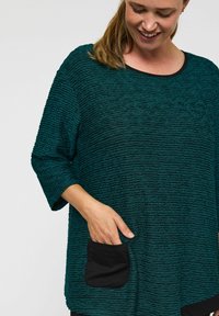 Zeleni teksturirani top z udobnim krojem, okroglo ovratnikom in tri četrtinskimi rokavi. Ima črn kontrastni žep na sprednji strani.