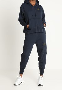 Persona con sudadera oscura de color azul marino y pantalones jogger con logos dorados de Nike, y zapatillas blancas de Nike, de pie con las manos en el bolsillo de la sudadera.