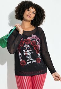 Sort mesh langærmet skjorte med et farverigt Day of the Dead kraniumgrafik omgivet af roser, parret med pinkstribede leggings og en grøn taske.