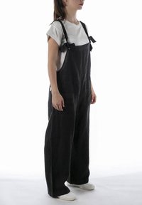 Salopette nera in lino con design a gamba larga, spalline regolabili da annodare e tasche frontali. Indossata sopra una camicia bianca a maniche corte.