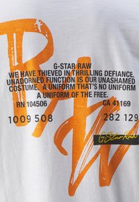 Camiseta blanca con un gran gráfico naranja y texto negro. Presenta la marca G-Star Raw y varios códigos numéricos. Material de algodón suave.