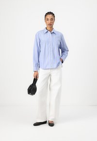 Blue and white striped button-up shirt with a collar, paired with white wide-leg trousers and black flats, holding a black handbag.