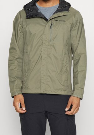 Man die een olijfgroene Columbia-jas draagt met een zwarte capuchon en zwarte broek, staand met handen langs zijn zij tegen een witte achtergrond.