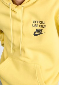Žlutá mikina s kapucí, přední kapsou, stahovacími šňůrkami a černými logy "POUZE PRO OFICIÁLNÍ POUŽITÍ" a "NIKE". Měkká, strukturovaná látka.