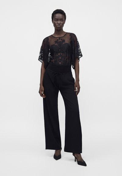 Femme portant un haut en dentelle florale noire transparente, un pantalon noir taille haute à jambes larges avec cordon, et des escarpins noirs pointus sur fond blanc.