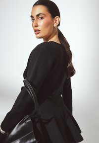 Maglione nero con corpino aderente e orlo a peplo svasato, abbinato a una borsa nera. Texture liscia e maniche lunghe visibili.