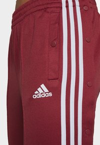 Bordeauxfarbene Sporthosen mit weißen Seitenstreifen, Druckknöpfen und einem Adidas-Logo. Gefertigt aus glattem Stoff mit einem elastischen Bund.