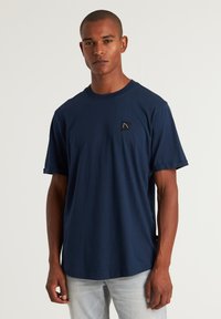 T-shirt in cotone navy con colletto girocollo, maniche corte e un piccolo patch quadrato del logo sul lato sinistro del petto. Vestibilità rilassata, texture liscia.