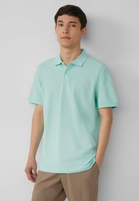 Polo shirt vert menthe clair avec un col, parement à deux boutons, manches courtes et un logo subtil sur la poitrine. Porté avec un pantalon beige.