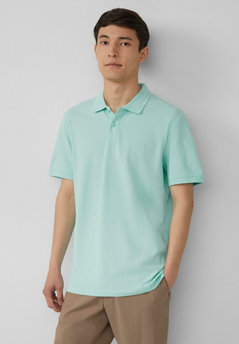 Polo shirt vert menthe clair avec un col, parement à deux boutons, manches courtes et un logo subtil sur la poitrine. Porté avec un pantalon beige.