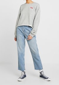 Grauer verkürzter Pullover mit rotem Markenlogo, kombiniert mit hellblauen Jeans mit geradem Bein und schwarzen High-Top-Sneakern.