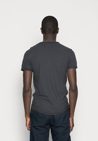 Hombre de pie de espaldas, vistiendo una camiseta de manga corta gris oscuro y pantalones azul marino, contra un fondo claro y sencillo.