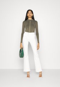 Top lung cu mânecă lungă de culoare verde măsliniu, pantaloni albi evazați, sandale cu toc transparent, ținând o geantă texturată verde, fundal simplu.