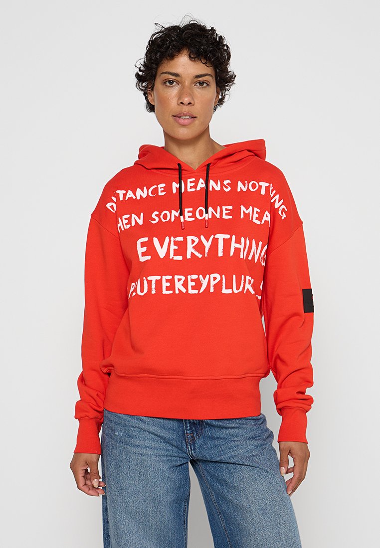 Osoba ubrana w czerwoną bluzę z kapturem z białym napisem "Dystans nic nie znaczy, gdy ktoś znaczy wszystko", w połączeniu z niebieskimi dżinsami.