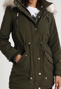 Kobieta w oliwkowej parkowej kurtce z futrzanym kapturem, zamkiem błyskawicznym i zatrzaskami, z rękami w przednich kieszeniach.