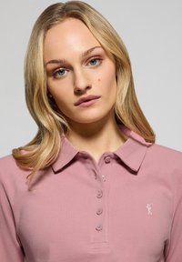 Ljusrosa polo skjorta med krage, utrustad med en placket med knappar och en liten broderad logotyp på bröstet. Slät, texturerad tyg.
