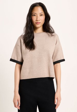 Woman with long dark hair wearing a beige short-sleeve top with black trim and black high-waist pants against a plain background.