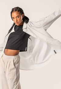 White button-up shirt layered over a black cropped top, paired with light beige pleated trousers. Smooth textures and casual fit.