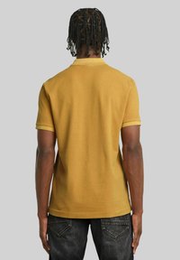 Polo shirt giallo senape a maniche corte con colletto piatto e polsini a contrasto, abbinato a jeans di denim scuro. Tessuto di cotone liscio.