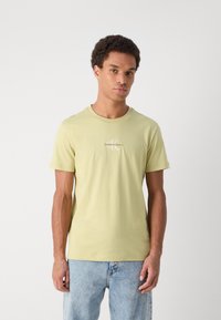 Gai zaļš kokvilnas T-krekls ar īsām piedurknēm, ar mazu balto "Calvin Klein Jeans" logo uz krūtīm, apaļā kakla dizains.