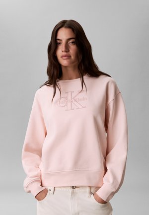 Γυναίκα με μακριά καστανά μαλλιά που φοράει ανοιχτό ροζ φούτερ Calvin Klein και λευκό παντελόνι, στέκεται μπροστά σε απλό γκρι φόντο.