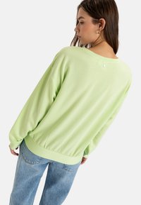 Light green loose-fitting sweatshirt with long sleeves, elastic hem, and a simple button detail at the back. Paired with blue jeans.