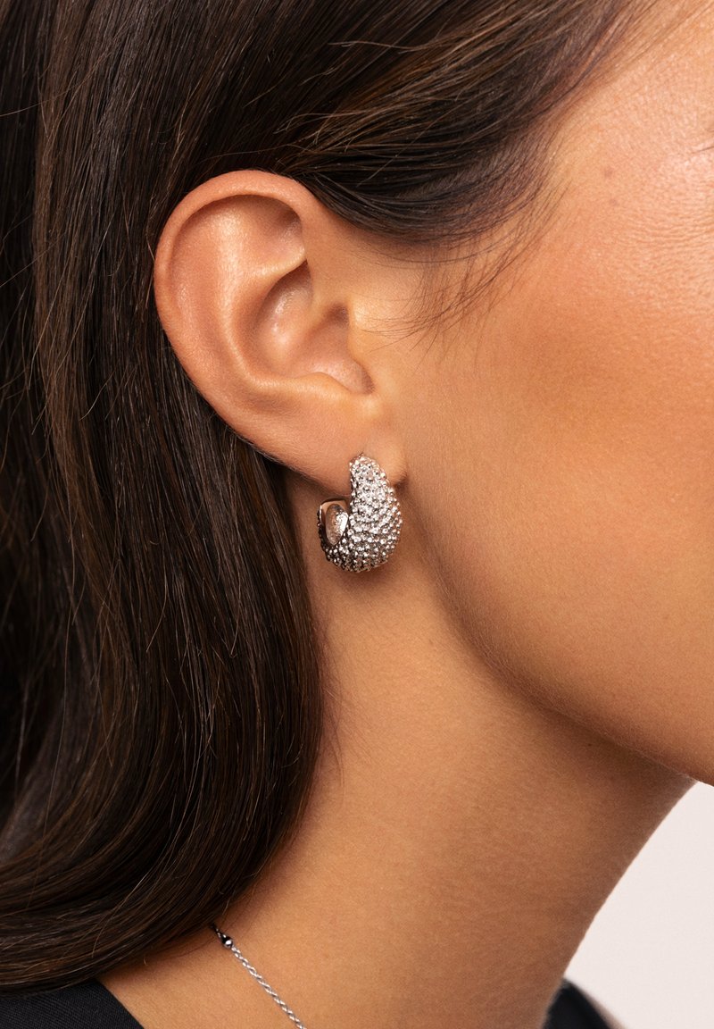 Srebrne kolczyki o zakrzywionym kształcie, ozdobione błyszczącymi kryształowymi akcentami i polerowanym wykończeniem. Prezentowane na uchu w widoku z profilu.