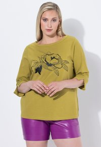 Top verde oliva con cuello redondo, mangas de tres cuartos y diseño floral negro. Combinado con pantalones cortos morados brillantes. Textura de tela suave.