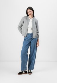 Grå bomberjakke, hvit skjorte, løse blå jeans og svarte sneakers med snøring. Myk stoff, ribbede mansjetter og en avslappet passform.