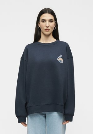 Kvinde med langt mørkt hår iført store øreringe, en navy oversized sweatshirt med lille grafik på brystet og lyseblå jeans.