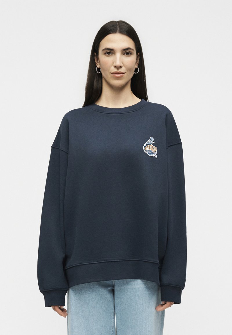 Femme aux longs cheveux foncés portant des boucles d'oreilles anneaux, un sweat-shirt oversize bleu marine avec un petit motif sur la poitrine et un jean bleu clair.