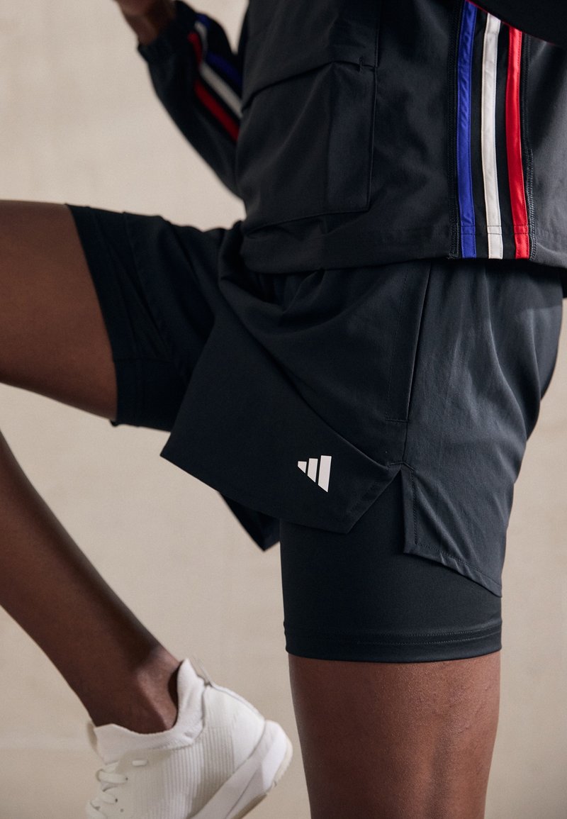 Personne portant un short de sport noir et une veste avec des rayures rouges, blanches et bleues, levant la jambe, et une basket blanche au pied.