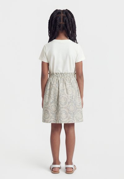 Κορίτσι με πλεξούδες στέκεται με την πλάτη γυρισμένη, φορώντας άσπρο κοντομάνικο τοπ, φούστα με σχέδια και ελαστική μέση, και άσπρα σανδάλια.