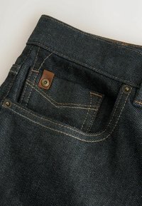 Sötét farmernadrág hátul zsebbel, amely bőrkiegészítéssel, fém szegecsekkel és kontrasztos varrással van díszítve a részletek kiemeléséhez.